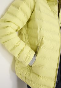 Doudoune jaune clair avec texture matelassée, fermeture éclair à l'avant et poche latérale. Poignet de pull gris visible à l'intérieur de la veste.
