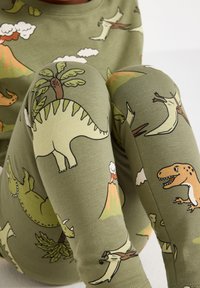 Bambino che indossa leggings verdi con fantasie colorate di dinosauri e vulcani, seduto con le ginocchia piegate su uno sfondo bianco.