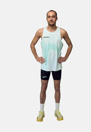 Mężczyzna stoi z rękami na biodrach, ubrany w jasnoniebieski tank top Brooks, czarne spodenki, białe skarpetki i żółte buty do biegania.
