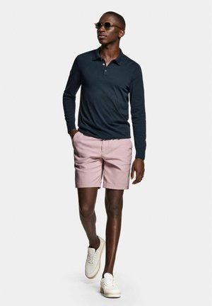 Marineblauw poloshirt met lange mouwen, roze shorts en witte sneakers. Zonnebril op. Gepolijste stof, gestructureerd ontwerp en contrast tussen kleuren.
