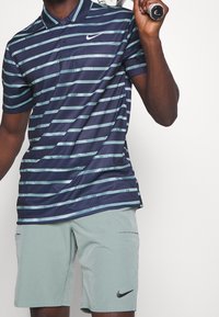 Chemise polo Nike avec des rayures horizontales bleues et bleu-vert clair, dotée d'un col et de manches courtes. Associée à un short vert clair, également de Nike.