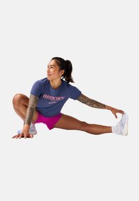 Ropa deportiva que incluye una camiseta azul con el logo "BROOKS", pantalones cortos rosas y zapatillas blancas. El modelo está estirándose en una pose dinámica.