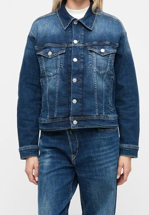 Personne portant une veste en jean bleue avec des boutons à l'avant et deux poches à rabat sur la poitrine, assortie à un jean en denim bleu.