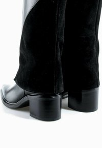 Bottes noires mi-mollet à talons blocs avec un pied en cuir lisse et une partie supérieure en suédine, vues de dos sur fond blanc.