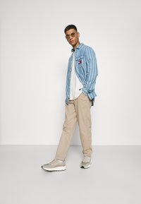 Blue striped button-up shirt, white t-shirt, beige pants, and grey sneakers; accessories include light-colored sunglasses.