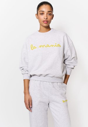 Nő áll, egyik kezét a zsebében tartja, világosszürke pulóvert és nadrágot visel, mindkettő elején sárga "la mania" felirattal.