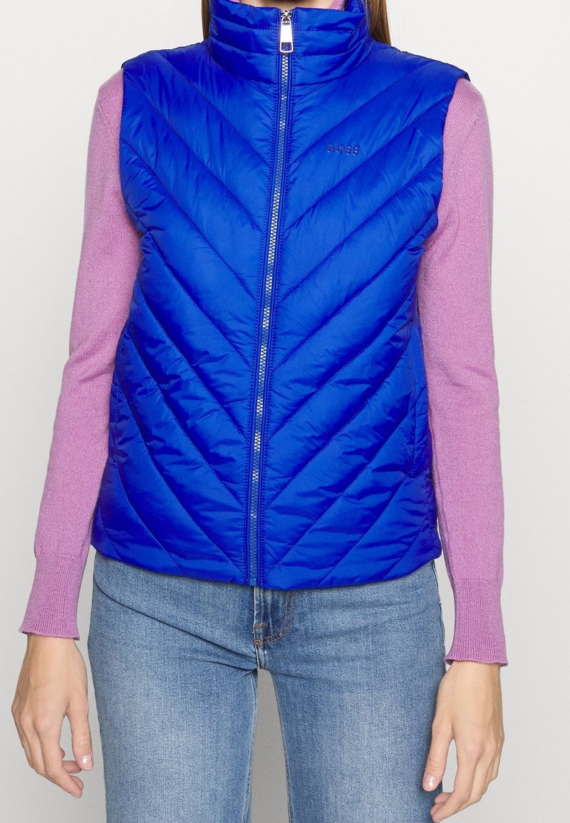 Blue quilted vest with a high collar, featuring chevron stitching and a front zipper. Worn over a pink long-sleeve top.