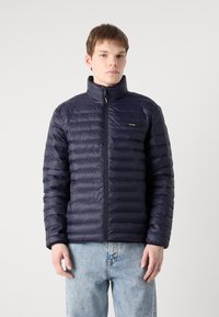 Navy puffer jacket featuring a zip closure, high collar, and horizontal quilted texture. Includes a chest logo patch and elastic cuffs.