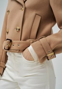 Abrigo de mezcla de lana beige de doble botonadura con grandes botones, cintura ceñida con cinturón y detalles en tono dorado, combinado con jeans blancos de cintura alta.