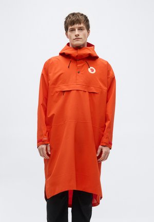 Mann trägt lange orangefarbene wasserdichte Kapuzenjacke mit Frontklappentasche und schwarze Hose, steht vor weißem Hintergrund.