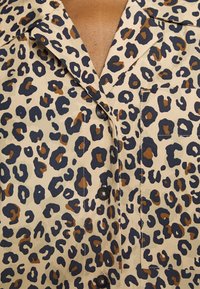 Camisa bege com padrão de leopardo em castanho escuro e azul marinho. Apresenta um colarinho em V, fecho frontal com botões e um bolso de patch. Tecido texturizado.