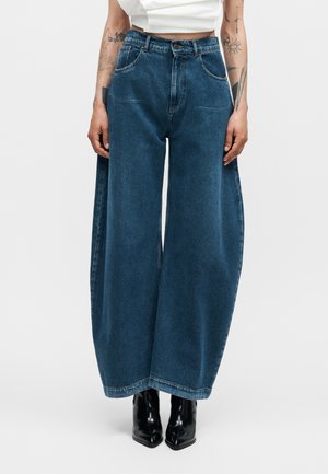 Jean denim bleu à jambes larges porté avec des bottes noires à bout pointu et un top court blanc, bras tatoués visibles sur les côtés.