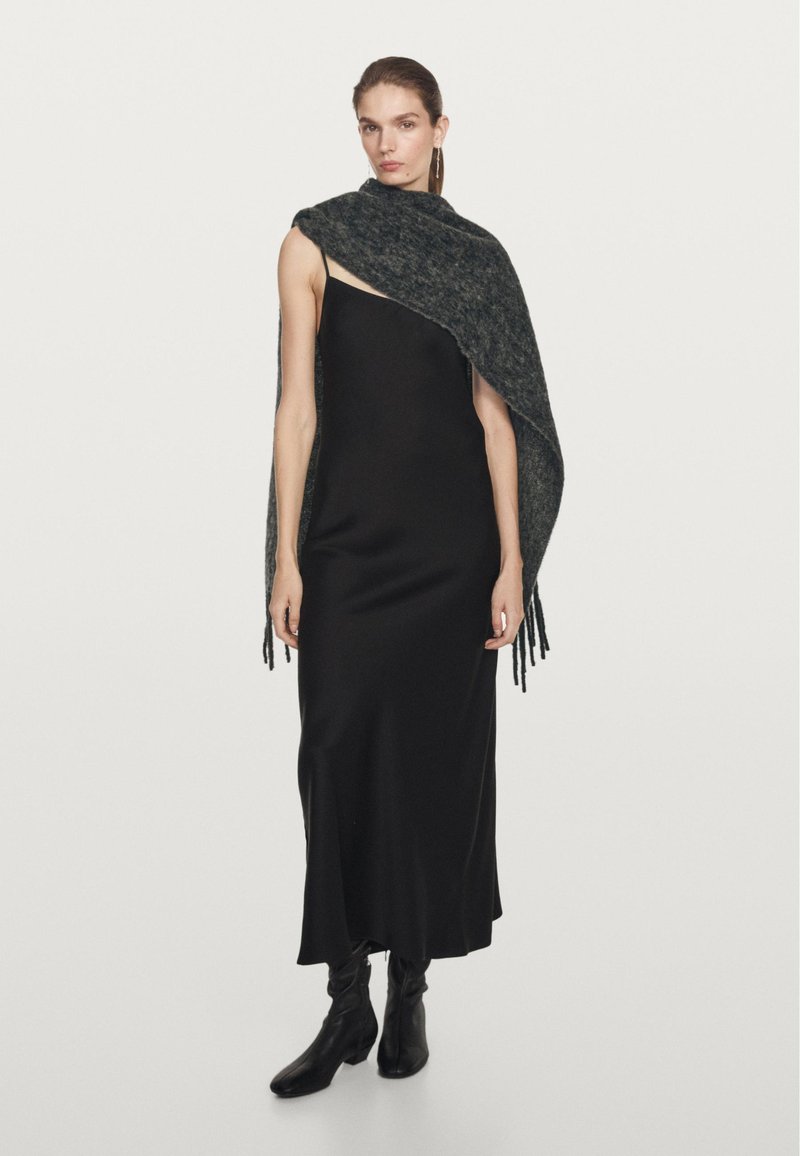 Černé saténové maxi šaty v kombinaci s tmavým texturovaným šálem se třásněmi. Model má na sobě černé kotníkové boty a jednoduché naušnice.