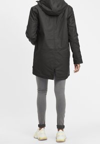 Czarna wodoodporna kurtka z kapturem, sięgająca do połowy uda, z tylnym wentylkiem i gładką teksturą; noszona na szarych dżinsach i sneakersach.