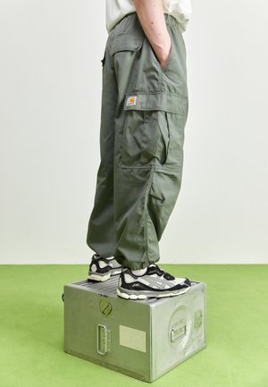 Personne portant un pantalon cargo vert et des baskets noir et blanc, debout sur une boîte en métal argentée sur un sol vert.