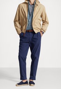 Béžová bomber bunda, světlá modrá denimová košile se pruhy, tmavě modré kalhoty, hnědý pletený pásek, tmavě modré espadrilky a hodinky na zápěstí.