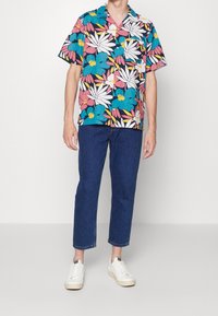 Short-sleeve button-up shirt featuring a vibrant floral pattern with blue, pink, and white colors. Paired with denim jeans and white sneakers.