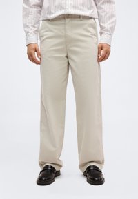 Άνδρας που φοράει μπεζ παντελ όνι με φαρδιά μπατζάκια, ροζ ριγέ πουκάμισο που είναι κομμένο μέσα, και μαύρα δερμάτινα παπούτσια στέκεται σε λευκό φόντο.