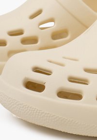 Zoccoli leggeri beige con una superficie liscia, caratterizzati da diversi fori di ventilazione e un design sagomato per il massimo comfort.