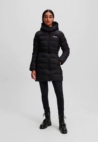 Abrigo acolchado negro con capucha, cremallera frontal, cintura con cinturón y bolsillos laterales. Acabado brillante con textura acolchada, combinado con pantalones y botas negras.