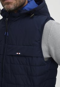 Tamsiai mėlynas pūkinis liemenės modelis su kapučiu, su pilkais akcentais, siūtomis panelėmis ir mažo logotipo detale raudonos, baltos ir mėlynos spalvų.