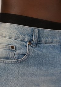 Jasnoniebieskie dżinsy z czarnym elastycznym pasem, z mosiężnymi detalami, delikatnym przetarciem i otworem kieszeni z przodu.