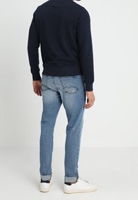 Pánská námořnická mikina s kulatým výstřihem, v kombinaci se světle modrými opotřebovanými džíny a bílými teniskami, ukazující neformální vzhled ze zadní strany.