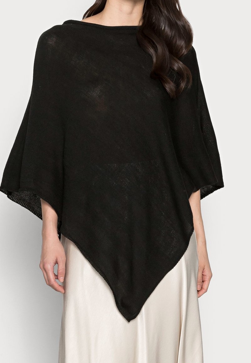 Femme portant un poncho en maille noire ample avec un ourlet pointu sur une jupe de couleur claire, bras le long du corps, sur un fond uni.