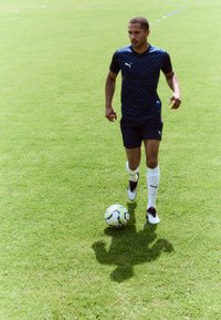 Plava majica s uzorkom za sport, tamnoplave šortse, bijele čarape i crne kopačke. Nogometna lopta na travi, s sjenom igrača koja je vidljiva.