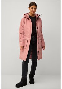 Roze waterdichte lange jas met capuchon, met rits en knoopsluiting aan de voorkant, twee zakken aan de voorkant, gedragen over een zwart shirt en donkere spijkerbroek, met zwarte laarzen.