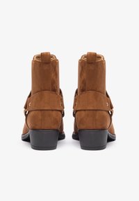 Brune semskede ankelstøvler med en glat tekstur, der har en dekorativ rem og en lille blokhæl. Designet er enkelt og kompakt.