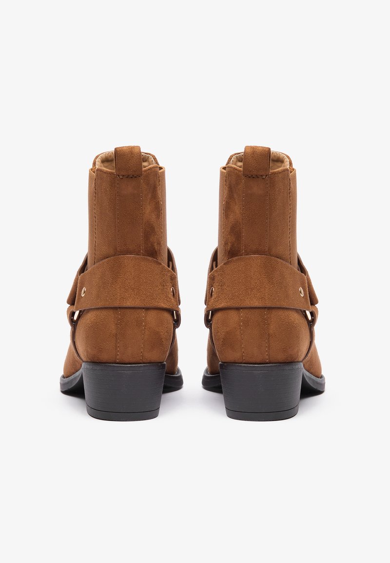 Botas de tobillo de ante marrón con una textura suave, que cuentan con una correa decorativa y un pequeño tacón cuadrado. El diseño es simple y compacto.