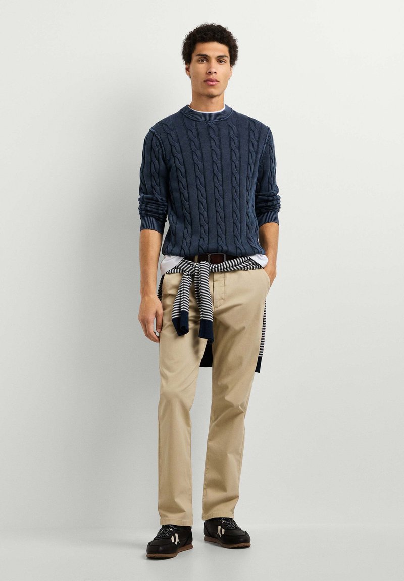 Jeune homme portant un pull en maille torsadée bleu marine et un pantalon beige, avec un pull rayé noué autour de la taille, debout contre un mur clair uni.