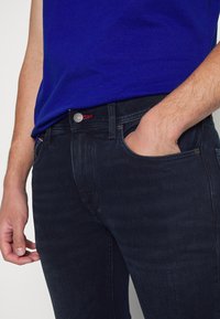 Jeans de mezclilla azul oscuro con un ajuste elegante, que presentan un botón plateado, detalles de costura en rojo y bolsillos laterales. Textura suave.