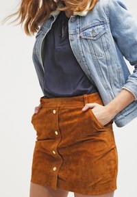 Mini-jupe en daim marron avec boutons sur le devant, poches latérales et ourlet légèrement incurvé, associée à une veste en jean bleue et un haut foncé.