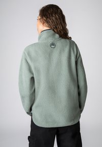 Lichtgroene fleecetrui met een gestructureerd oppervlak, hoge kraag en circulair logo aan de achterkant, gecombineerd met zwarte cargobroek.