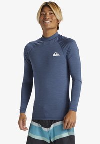 Tamno plavi rash guard za surfanje s dugim rukavima i visokim ovratnikom. Ima mali bijeli logo na grudima i glatku teksturu tkanine.
