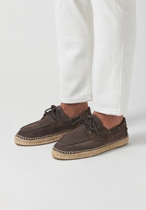 Braune Wildleder-Espadrilles mit einer gewebten Jutesohle, ausgestattet mit Schnürsenkeln und metallischen Ösen. Getragen mit weißen Hosen, die das Design des Schuhs hervorheben.