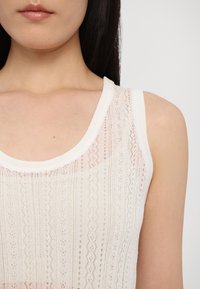 Gaiši bežs bez piedurknēm topiņš ar mežģīņu līdzīgu tekstūru un vertikālām rakstu līnijām. Apaļš izgriezums.