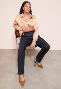 Satin beige skjortan med knappar, mörka jeans, och bruna spetsiga klackar, sittande på en metallstol mot en neutral bakgrund.