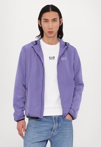 Casaco lilás claro com capuz, fecho de correr à frente e punhos elásticos, usado sobre uma t-shirt branca com um pequeno logotipo. Calças de ganga casuais.
