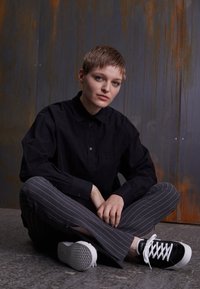 Camicia nera con bottoni, pantaloni grigi a righe e sneaker nere con lacci bianchi. Persona seduta a terra, con uno sfondo testurizzato.