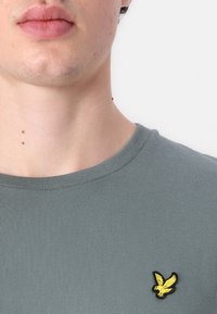 Camiseta gris de rayas con cuello redondo, que presenta un prominente logotipo de águila bordado en negro y amarillo en el lado izquierdo del pecho.