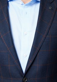 Granatowa marynarka w kratę z brązowymi liniami, z czarnym guzikiem i klapami, nałożona na jasnoniebieską koszulę z kołnierzykiem z białymi guzikami i teksturowanym materiałem.