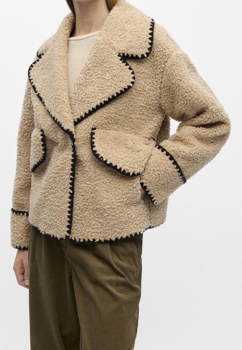 Veste en polaire beige texturée avec une bordure zigzag noire sur le col, les poches, les poignets et l'ourlet, portée sur une chemise claire et un pantalon vert olive.