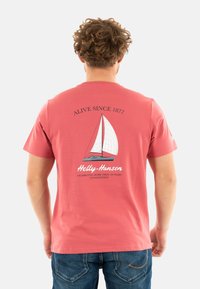 Camiseta de manga corta de color coral con un gráfico de un bote de vela, con el texto "VIVO DESDE 1877" y "Helly-Hansen" en la parte posterior.