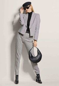 Grauer strukturierter Blazer und Hosenanzug mit einem taillierten schwarzen Rollkragenpullover. Das Model hält eine schwarze geschwungene Handtasche und trägt schwarze Sonnenbrille und eine Mütze.