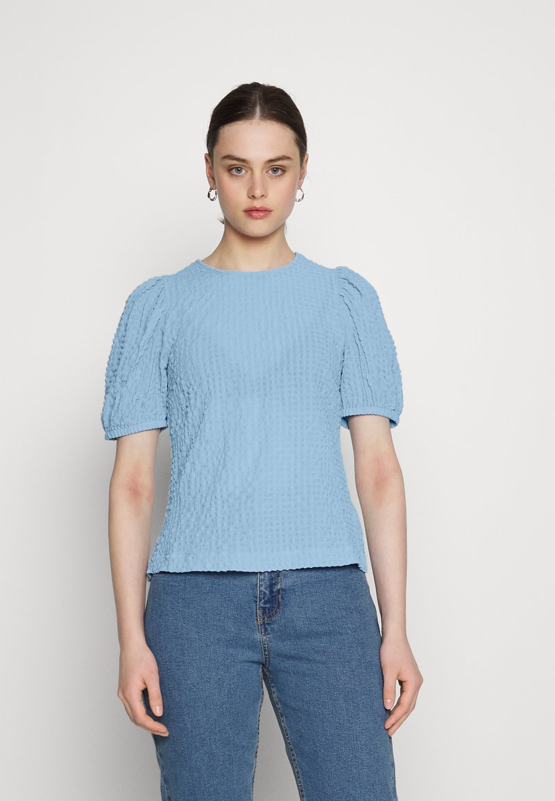 Blusa texturizada azul clara com mangas curtas em balão e decote redondo. Usada com jeans de cintura alta. Design minimalista.