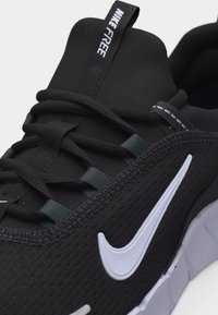 Κοντινό πλάνο ενός μαύρου αθλητικού παπουτσιού Nike Free με λευκό λογότυπο Swoosh, μαύρα κορδόνια και μια θηλιά έλξης με την ένδειξη "Nike Free" στη γλώσσα.