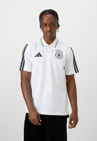 Camisa polo branca com riscas pretas nas mangas, apresentando o logo preto da Adidas e um emblema da Alemanha com quatro estrelas. Design de mangas curtas e colarinho.